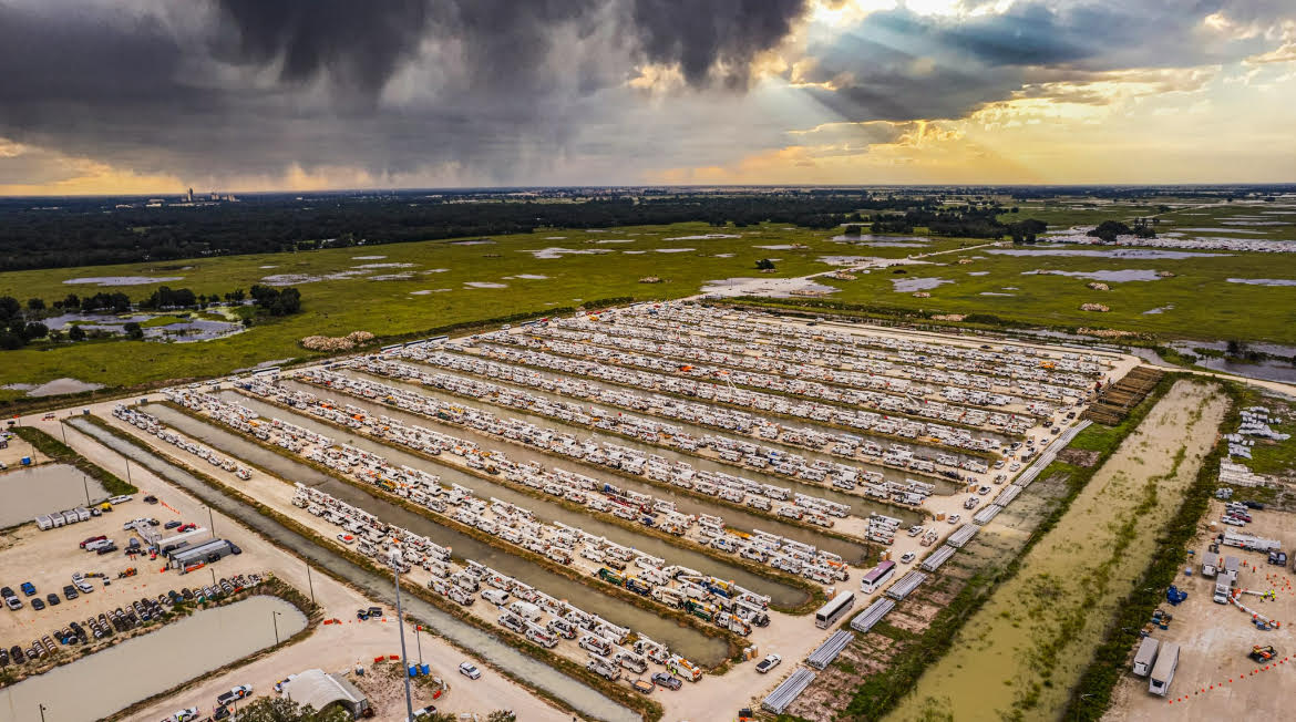 Aerial view of a utility contractor staging area — hundreds of bucket trucks lined up under a storm sky